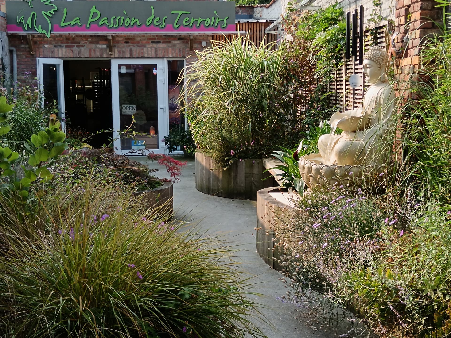 L’entrée de La Passion des Terroirs : allée fleurie bordée de végétation menant à la façade en briques